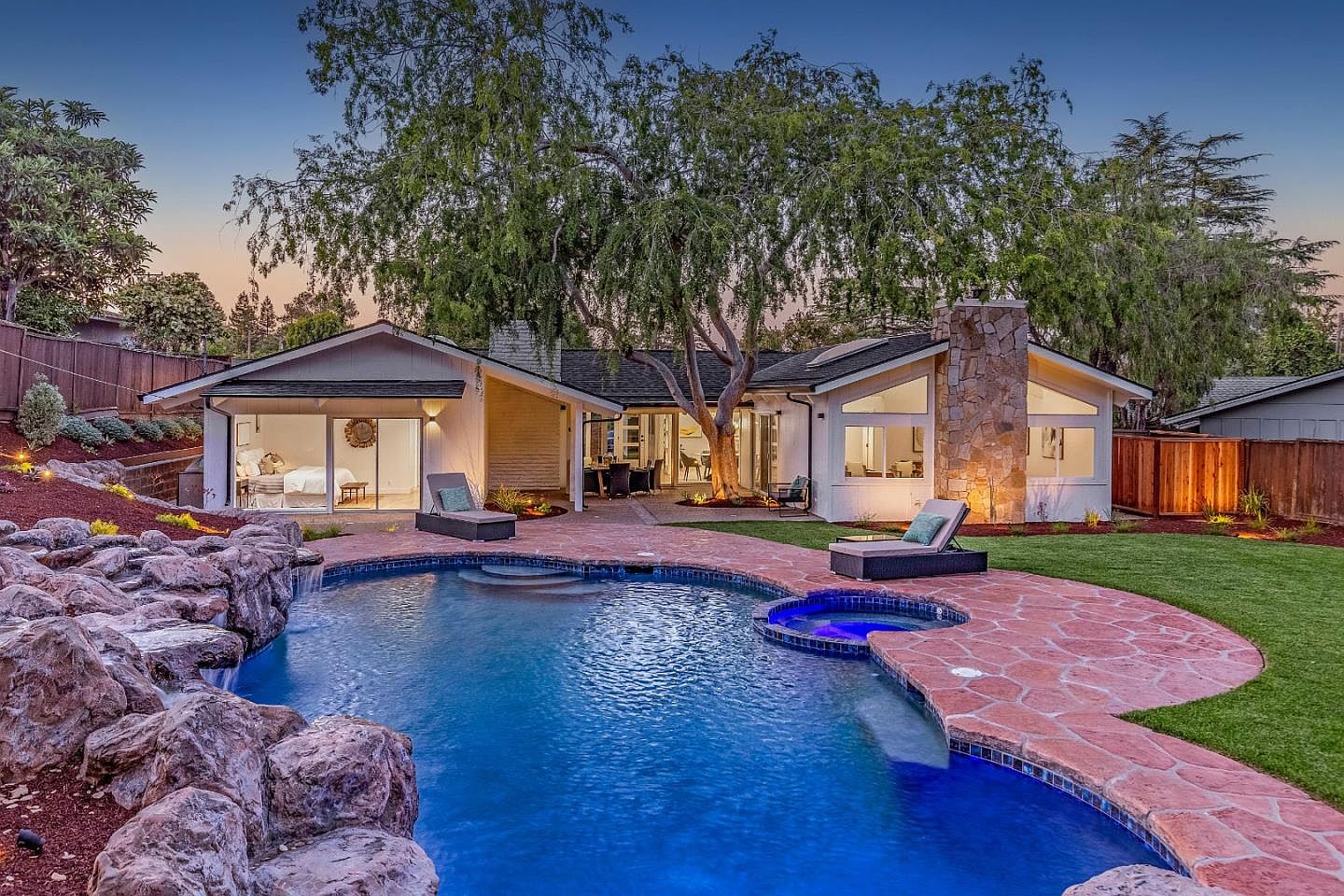 The backyard at 1677 Middleton at twilight, with a freeform pool and raised spa, flagstone decking, lounge chairs, and the rear of the house glowing through picture windows