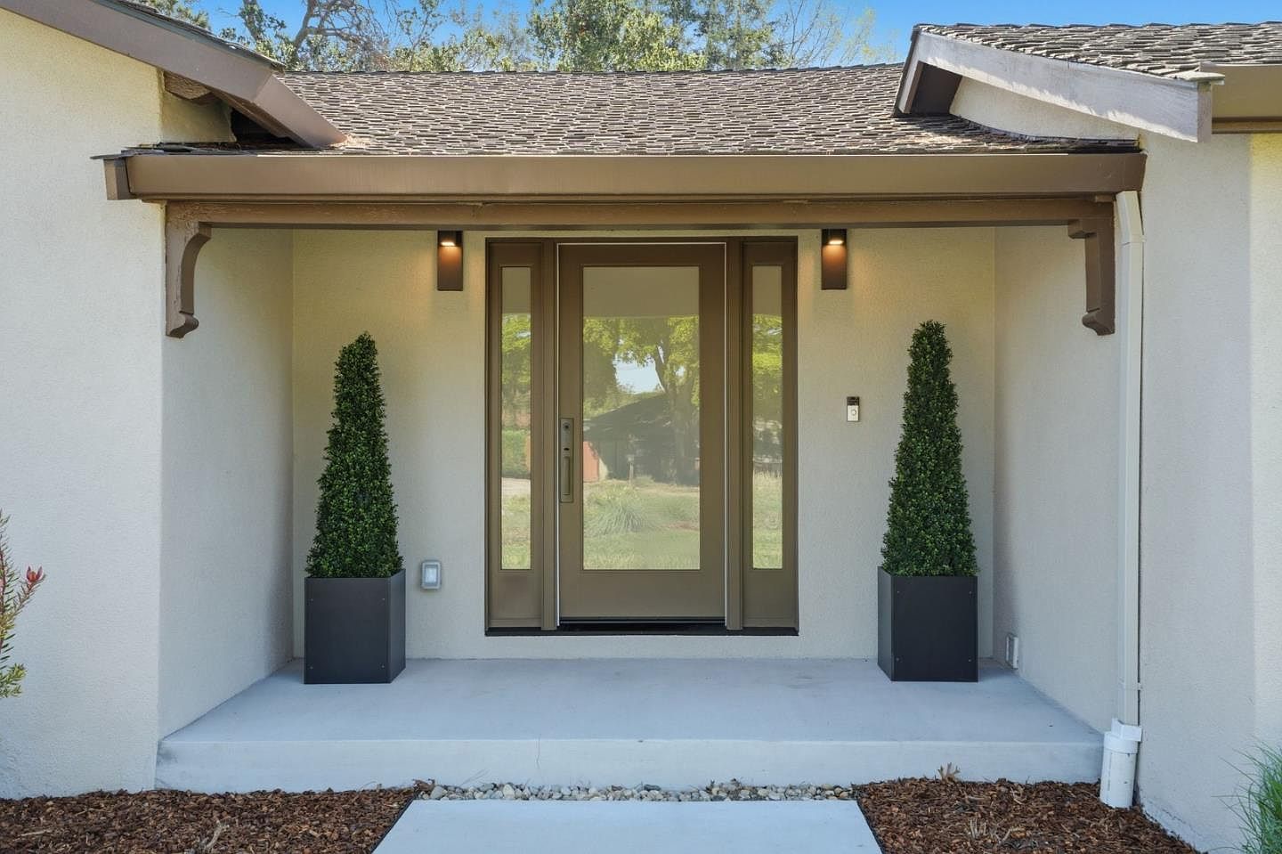 The covered front entry of 1677 Middleton with a glass-paneled door flanked by sidelights, deep eaves, and two clipped topiaries on a poured-concrete porch