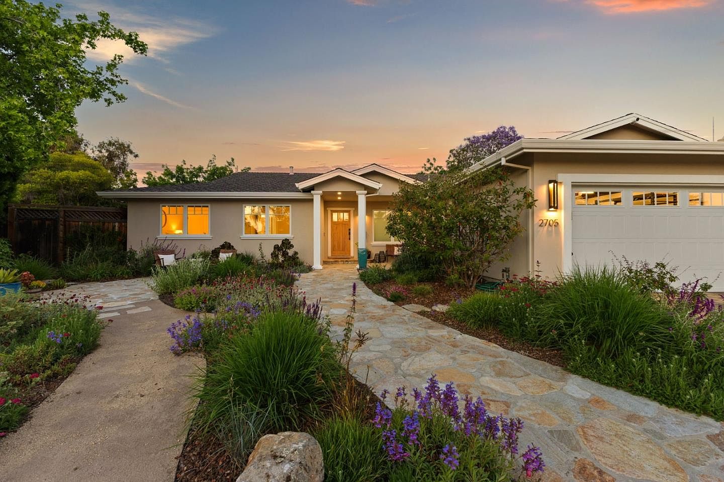 Front elevation of 1677 Middleton at twilight, the pale stucco facade and warm-lit windows glowing against a pink dusk sky above a flagstone path through the native garden