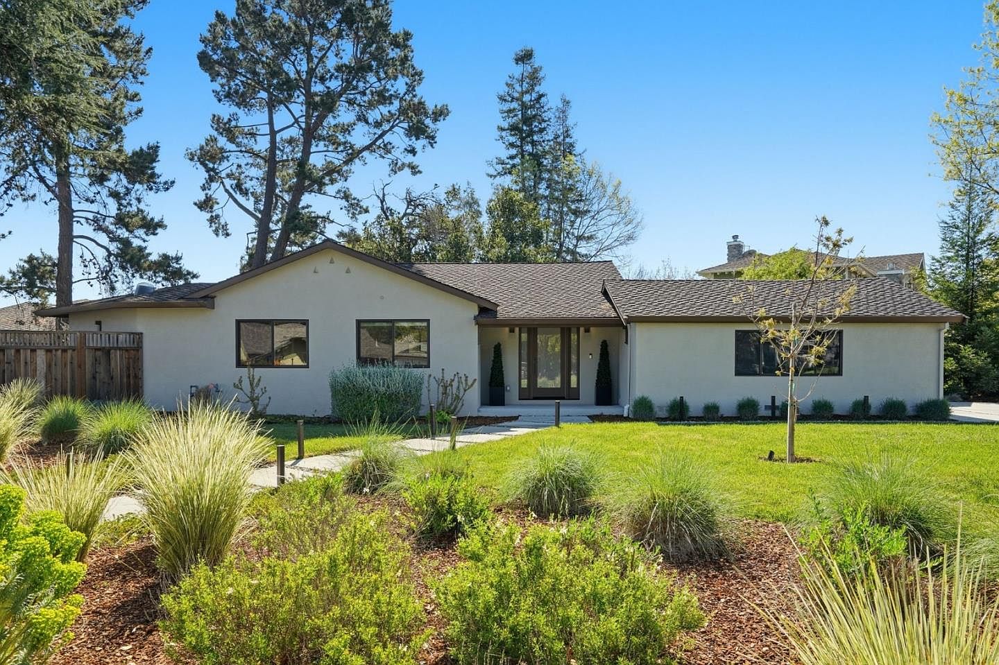 Wide front view of 1677 Middleton across the lawn, showing the full single-level footprint, the new roof, dark-framed windows, and mature pines along the property line